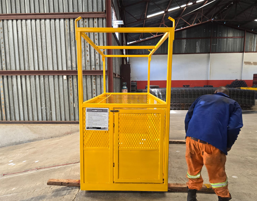 Yellow fork-mounted forklift safety cage with mesh sides, a gate and a data plate, standing in a warehouse